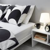 Bett mit BRUKSVARA schwarz/weißen Bogenkissen, weißer Lampe, Becher, Uhr und Büchern auf dem Nachttisch.
