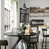 Modernes Café mit schwarzem Tisch, grauen Stühlen, Zirbengewürz-Organizer, Gläsern, Kreidetafelkarte und weißem Hintergrund.