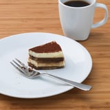 Eine Scheibe Schichtdessert mit einer Gabel auf einem weißen Teller neben einer Kaffeetasse.