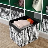 Schwarz-weiß gemusterte Aufbewahrungsbox auf dem Regal mit gefalteter Decke und orangefarbener Blume. Teil des Kallax Regals.