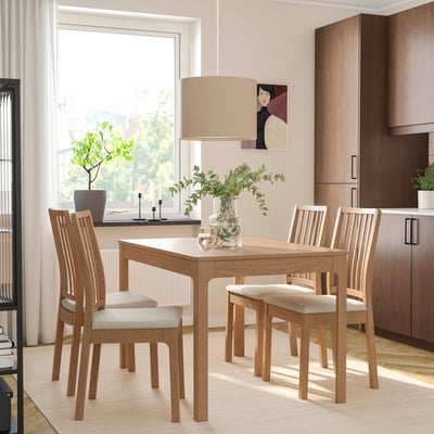 Esstisch aus Holz mit ausziehbarer Platte, gepolsterte Stühle. Natürliches Licht durch großes Fenster, Vase mit Grün, Kerzen.