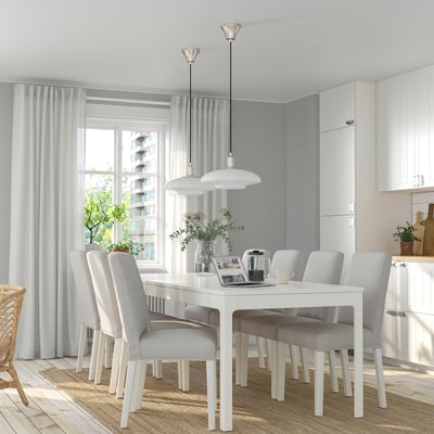 Moderne weiße Essgarnitur mit ausziehbarem Tisch und grauen Stühlen, großem Fenster mit Vorhängen, weißen Pendelleuchten, Topfpflanzen und Küchenschränken.