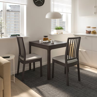 Moderne Küchenecke mit kleinem braunem EKEDALEN Tisch und Stühlen, weißen Schränken, großen Fenstern und einer hängenden weißen Lampe.