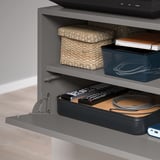 Das Bild zeigt einen modernen grauen EKET-Lagerraum. Das untere Regal verfügt über eine aufsteckbare Front für versteckte Aufbewahrung und Kabel, die für die Organisation verwaltet werden.