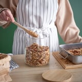 Eine Person schöpft Müsli aus einem durchsichtigen Glas auf einem Holztisch, ideal für die Aufbewahrung und den einfachen Zugriff auf trockene Lebensmittel.