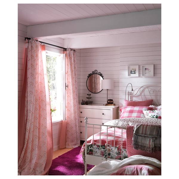 Gemütliches Schlafzimmer im rosa Stil mit weißen Möbeln, geblümter Bettwäsche, rosa Vorhängen und einem runden schwarzen dekorativen Spiegel an der Wand.