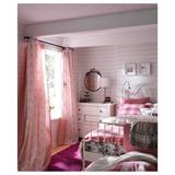 Gemütliches Schlafzimmer im rosa Stil mit weißen Möbeln, geblümter Bettwäsche, rosa Vorhängen und einem runden schwarzen dekorativen Spiegel an der Wand.