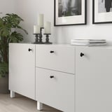 Weißer Schrank mit schwarzen Knöpfen, in dem Kerzen und Bücher stehen, neben einer Pflanze.