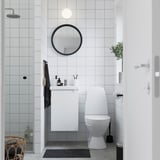 Modernes Badezimmer mit weißem ENHET-Schrank, rundem Spiegel, WC und Pflanzen. Das glänzende Gerät lässt sich hoch montieren und passt in enge Räume.