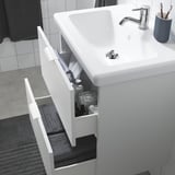 Weißer Badezimmerschrank mit Waschbecken und zwei offenen Schubladen. In einem befinden sich Toilettenartikel, in dem anderen ein Handtuch. Das Waschbecken verfügt über einen silbernen Wasserhahn.