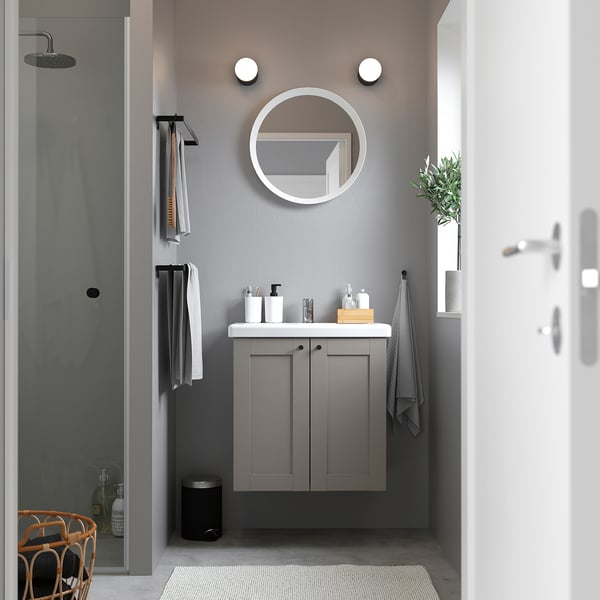 Modernes graues Badezimmer mit einem kleinen, an der Wand montierten ENHET-Waschtisch und einem runden Spiegel. Handtücher und Produkte ordentlich in Regalen organisiert. Dusche in der Ecke.