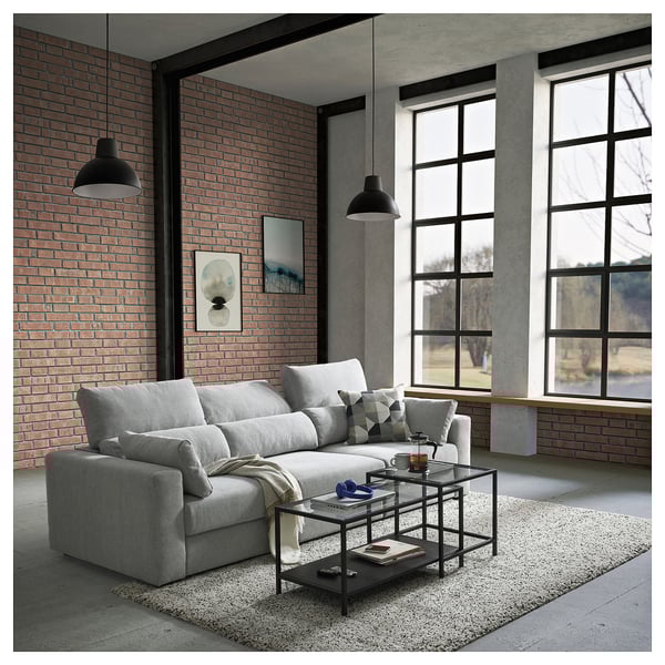 Ein modernes, graues ESKILSTUNA-Sektionssofa vor einer Backsteinwand, mit großen Fenstern, einem gläsernen Couchtisch und schwarzen Pendelleuchten.