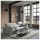 Ein modernes, graues ESKILSTUNA-Sektionssofa vor einer Backsteinwand, mit großen Fenstern, einem gläsernen Couchtisch und schwarzen Pendelleuchten.