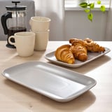 Tischdekoration mit drei Croissants auf rechteckigem Teller, zwei gestapelten Tassen und einer Kaffeepresse.