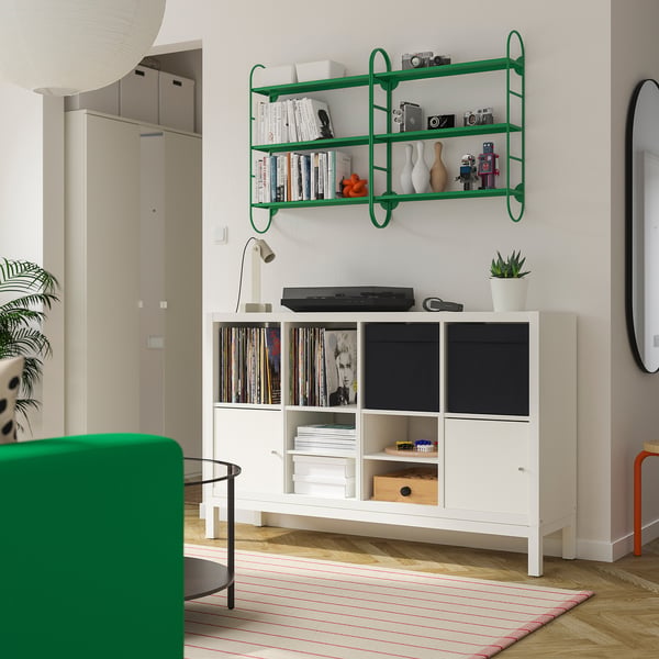 Grünes Regal mit Büchern, Vasen, Kameras über Schrank mit Schallplatte, Büchern, Drucker. Grüner Stuhl, gestreifter Teppich unten.