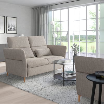 Modernes Wohnzimmer mit großem Fenster, grauem FAMMARP-Sofa, verstellbaren Kopfstützen, Holzbeinen, gemütlichem Teppich, schwarzem Couchtisch und Büchern.