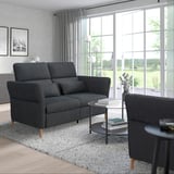 Modernes Wohnzimmer mit großem grauem FAMMARP-Sofa an der Wand, dunkelgrauem Teppich, Beistelltischen, Lampen und großen Fenstern.