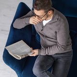 Mann sitzt auf blauem FAMMARP-Sofa, liest eine Zeitung, gekleidet in einen grauen Pullover und Jeans.