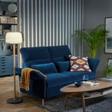 Blaues FAMMARP-Sofa mit verstellbaren Kissen, gemustertem Kissen, Beistelltisch aus Holz, Stehlampe und Pflanze.