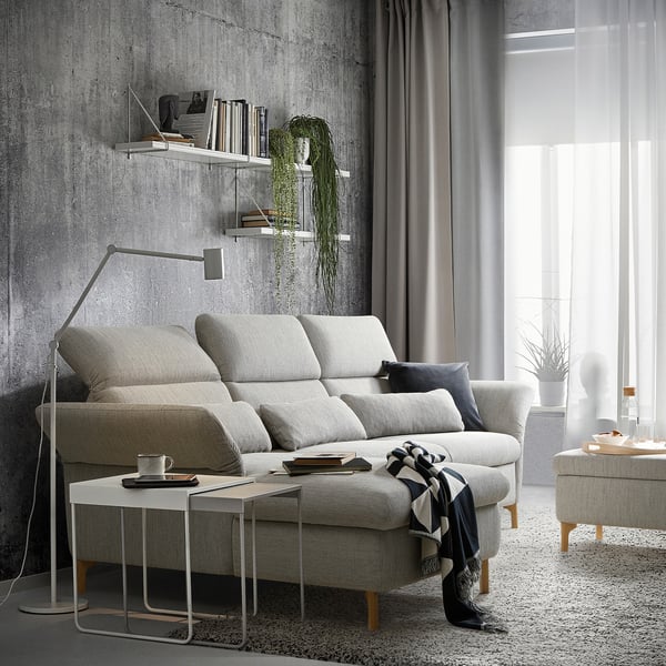 Modernes, minimalistisches Wohnzimmer mit grauem FAMMARP-Sofa, hölzernem Beistelltisch, Stehleuchte und Regalen an einer Betonwand, geschmückt mit Pflanzen und Büchern.