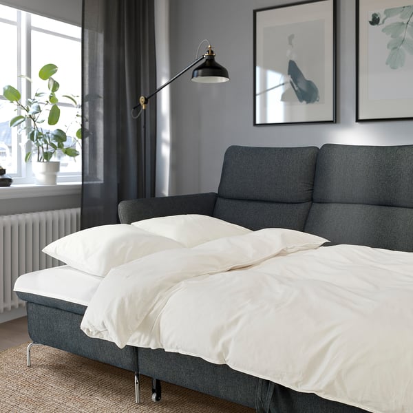 Modernes graues Schlafsofa mit weißer Bettwäsche, verstellbaren Kopfstützen und Armlehnen in einem gemütlichen Raum mit Wandkunst, Stehlampe und Pflanze.