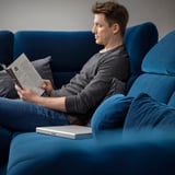 Mann sitzt auf blauem FAMMARP-Sofa und liest ein Buch mit einem geschlossenen Laptop auf der Armlehne. Das Sofa verstellbare Kopf- und Armlehnen.