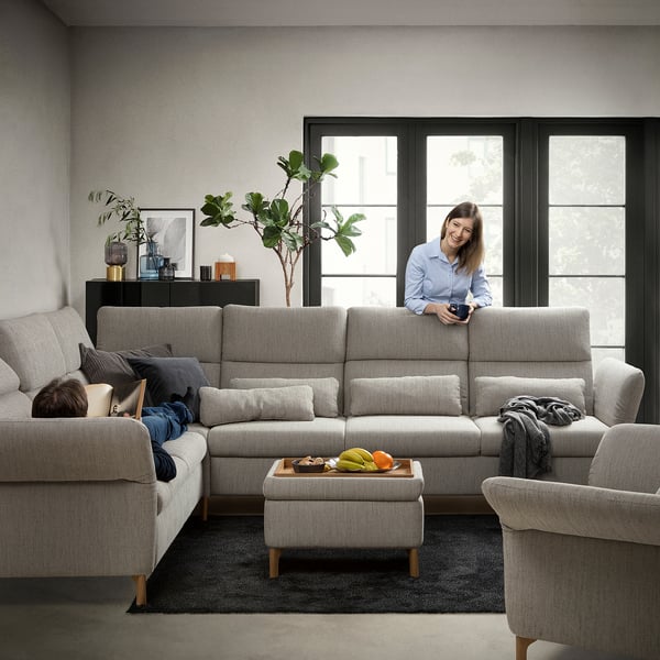Ein modernes Wohnzimmer mit einem hellgrauen FAMMARP-Sofa mit verstellbaren Kopfstützen und Armlehnen, einem schwarzen Teppich, großen Fenstern und einer Frau, die eine Fernbedienung in der Hand hält.