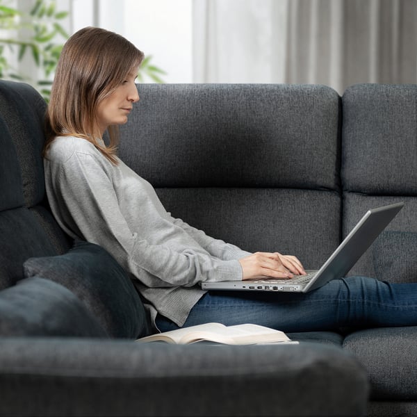 Eine Frau sitzt auf einem grauen FAMMARP-Sofa und benutzt einen Laptop, während neben ihr ein aufgeschlagenes Buch liegt.