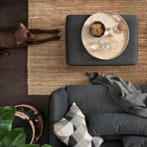 Eine Draufsicht auf eine dunkle Couch mit einem karierten Kissen, einer grauen Ottomane, einem Juteteppich und einer schlafenden Katze.