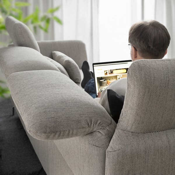 Älterer Mann mit Laptop auf verstellbarem FAMMARP-Sofa mit hochgelegten Füßen, grüner Pflanze, weißen Jalousien.
