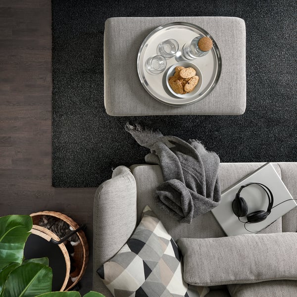 Draufsicht auf einen gemütlichen Wohnbereich mit einem grauen FAMMARP-Sofa, einem schwarzen Couchtisch, einer Pflanze und einem Holzkorb.