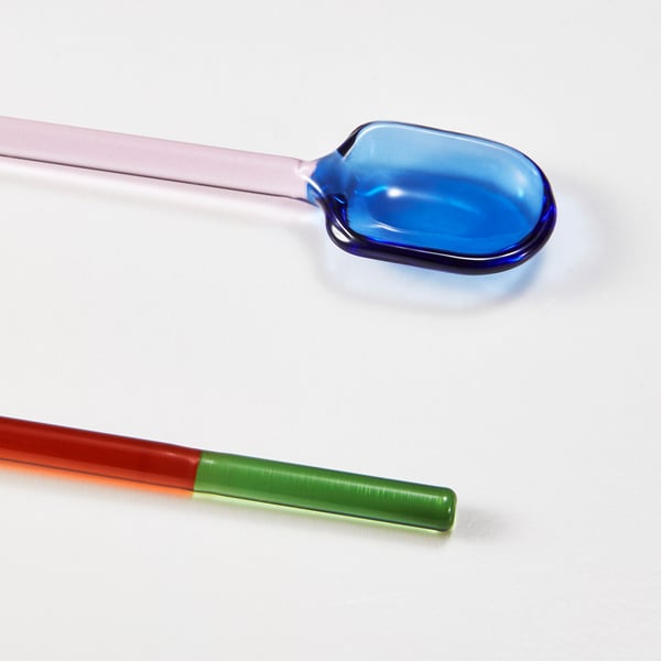 Zwei lebendige, bunte Glaslöffel auf weißer Oberfläche. Ein Löffel ist blau mit einem langen Stiel, der andere aus roten und grünen Segmenten.