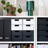 Moderner Schreibtisch mit FJÄDERHARV-Minitruhen in Weiß und Schwarz, kleiner Pflanze, grüner Lampe, Büchern und Dekor.