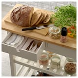 Ein hölzerner Küchenwagen mit Schubladen und Schränken, in dem Brot, Besteck und Lebensmittel ausgestellt sind.