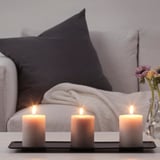 Drei brennende Kerzen auf einem Tablett mit einem Sofa und Blumen. Sorgt für Wärme und Ambiente.