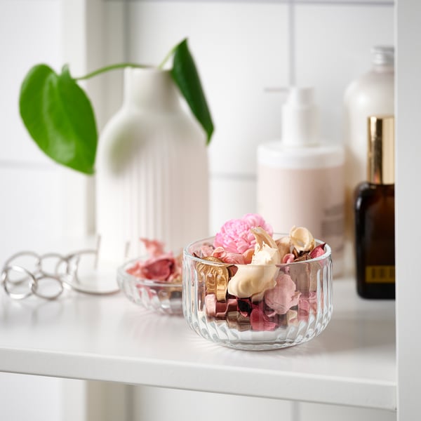 Eine kleine Glasschüssel gefüllt mit buntem Potpourri steht auf einem weißen Regal. Daneben eine weiße Vase mit einer grünen Pflanze und Schönheitsprodukten.