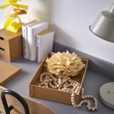 Schachteldisplay mit Holzperlen und getrockneten Blumen auf dem Schreibtisch, neben Büchern und Lampe.