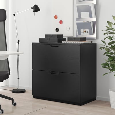 Schwarzer GALANT Aktenschrank mit 2 Schubladen neben weißem Schreibtisch. Tabletts auf der Oberseite nehmen Büromaterial auf. Schwarze Lampe und Stuhl vervollständigen minimalistisches Setup.