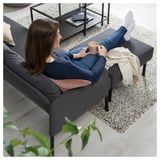 Eine Frau entspannt sich auf einem grauen GLOSTAD-Sofa und hält eine rosa Tasse in der Hand. Das Zimmer ist gemütlich mit einem gemusterten Teppich, einem Couchtisch und einer Pflanze neben dem Sofa.