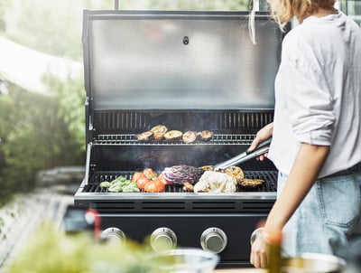 Person grillt Gemüse auf dem GRILLSKÄR-Grill im Freien mit einer Zange, mit Grün im Hintergrund.