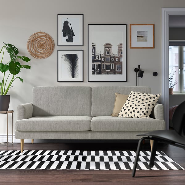 Modernes Wohnzimmer mit einem grauen GRILLSTORP-Sofa, einem schwarz-weißen Teppich und Wandkunst. Das Sofa hat klare Linien, Holzbeine und ist von Dekor umgeben.