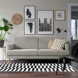 Modernes Wohnzimmer mit einem grauen GRILLSTORP-Sofa, einem schwarz-weißen Teppich und Wandkunst. Das Sofa hat klare Linien, Holzbeine und ist von Dekor umgeben.