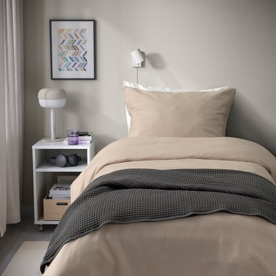 Beigefarbenes und graues Bettdeckenset mit Flanellstruktur. Twin-Size-Bett in der Nähe eines kleinen Tisches mit Büchern, Kopfhörern, Lampe. Bunter Wanddruck.