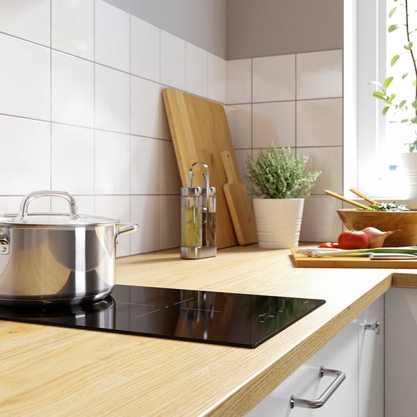Moderne Küchenarbeitsplatte mit schwarzem Induktionskochfeld, Holzschneidebrettern, Edelstahltopf, Zutaten und einer Topfpflanze.