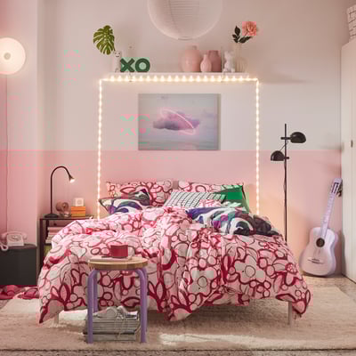 Rosa Schlafzimmer mit HÄRLIR-Bettdecke , weißen Wänden, Neon-Bettgestell, Hocker, Beistelltische, Gitarre, Vasen, XO-Schild, Wandposter.