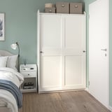 Modernes Schlafzimmer mit hohem weißem HAUGA-Kleiderschrank, Bett, Nachttisch, Lampe und Büchern. Hellblaue Wände, beruhigende Atmosphäre.