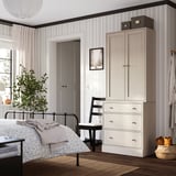 Gemütliches Schlafzimmer mit HAVSTA-Schrank, Truhe, Bett und Pflanze. Hochschrank mit Stuhl, Truhe mit Korb, sanfte Beleuchtung, großes Fenster.