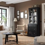 Schwarzer HAVSTA-Schrank mit Glastüren, Büchern. Daneben ein weißer Stuhl und ein schwarzer Couchtisch mit Vase. Zimmer mit Fotos, Lampe und orangefarbenem Teppich dekoriert.
