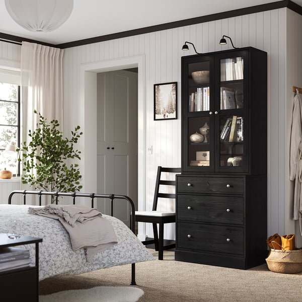 Modernes Schlafzimmer mit dunklen HAVSTA-Schubladen und Glasschrank, weißen Wänden, Metallbett, Pflanze, Stuhl, Teppich.