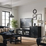 Modernes Wohnzimmer mit weißen Wänden, großem Fernseher auf dunklem Holzständer, blauer Couch mit Kissen, gestreiftem Teppich, Pendelleuchte, Pflanze und Dekorationsgegenständen.